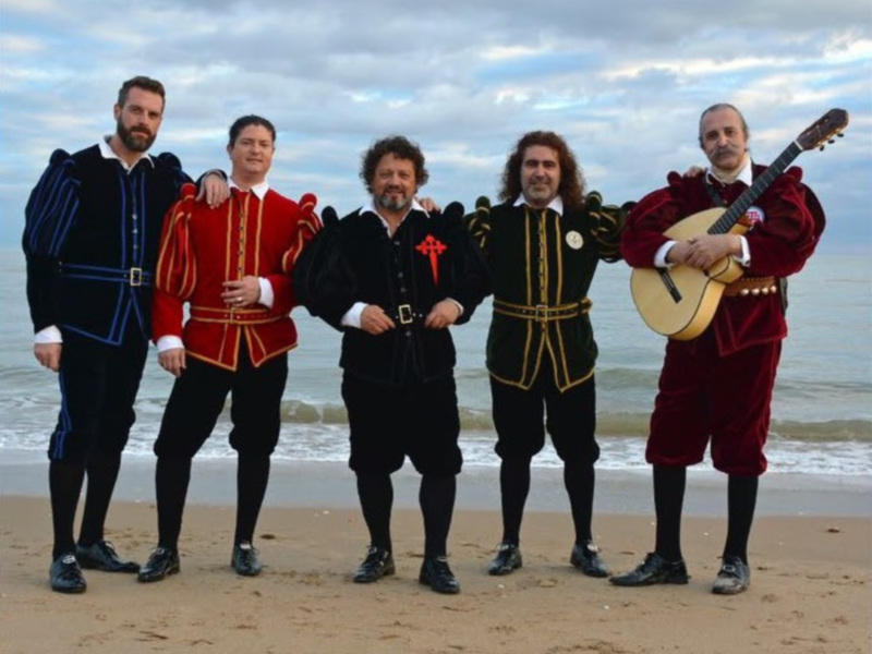Cuatro hombres vestidos con trajes medievales posan en la playa frente al mar, uno de ellos sostiene una guitarra medieval.
