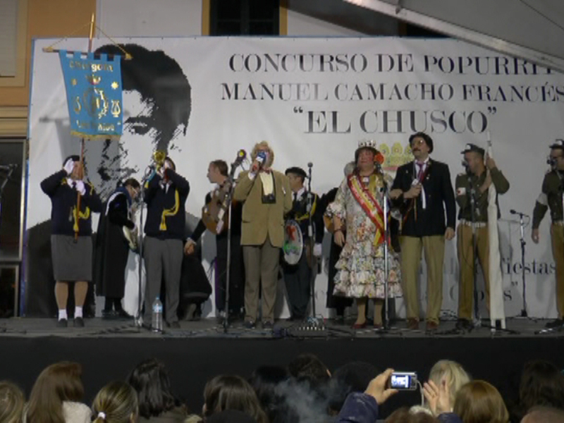 Una imagen de un grupo de personas en un escenario, con una pancarta que dice "Concurso de Popurrí". La gente está vestida formalmente y parece estar en un evento o ceremonia.