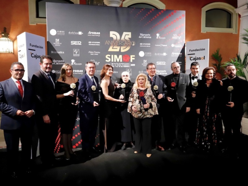 A una grupo de personas posando para una foto en un evento celebración del 25º aniversario. El fondo es un póster con logos de diferentes organizaciones y la palabra "25º ANIVERSARIO". La gente está vestida formalmente, con trajes y vestidos elegantes.