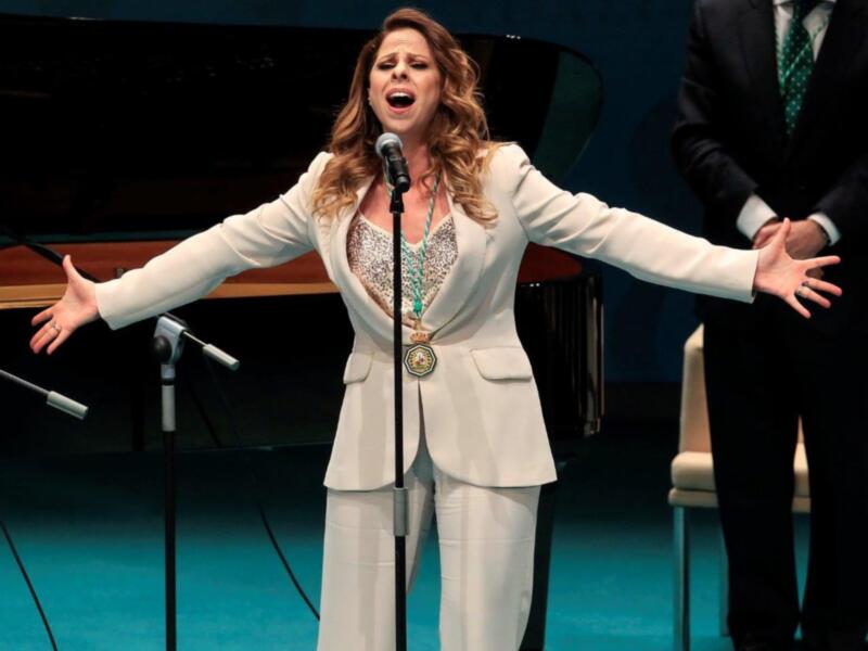 Cantante en actitud de canto sobre un escenario con piano y público en el fondo.