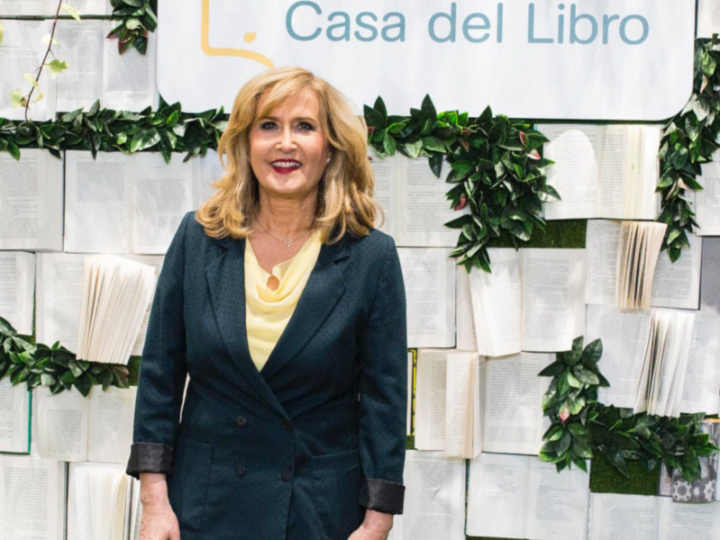Retrato de una mujer sonriente frente a un fondo decorado con libros y hojas verdes, en Casa del Libro.