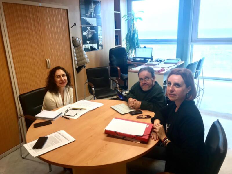 Tres personas sentadas en una mesa de oficina, con documentos y un teléfono móvil en el centro. La imagen muestra un ambiente laboral profesional con una ventana grande al fondo, iluminando el espacio.