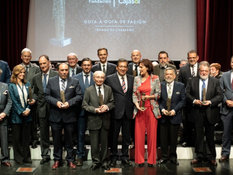 Una imagen de una gala de premiación donde un grupo de personas, principalmente hombres en trajes y mujeres en vestidos formales, se encuentran posando juntos. La escena está iluminada por luces de teatro y el fondo muestra un póster con texto que parece ser el nombre de la organización o evento. La celebración tiene un ambiente formal y festivo, indicando probablemente una ceremonia de reconocimiento o galardones.