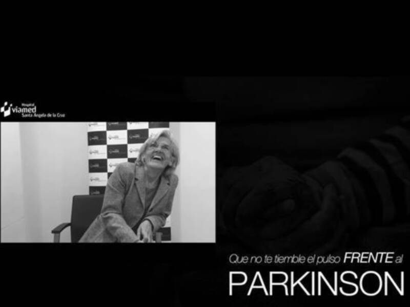 Una mujer sonriendo mientras se sienta en una silla, con un fondo que incluye un panel con logos y la palabra 'Parkinson'.