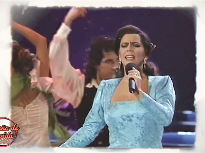 Cantante en escenario, vestida de azul, con micrófono en mano, rodeada de bailarinas en movimiento.