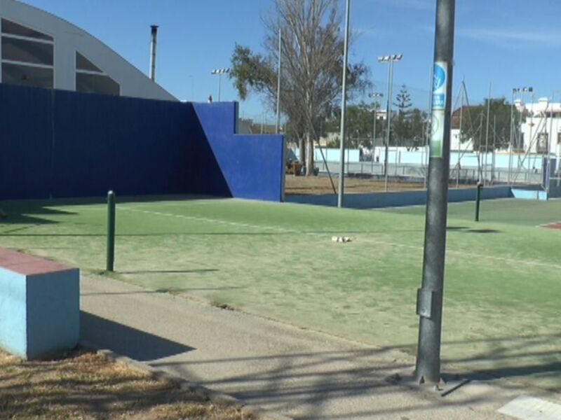 Cortina de seguridad azul en un campo deportivo con césped verde y una estructura de hormigón rojo y azul en el fondo. El cielo es azul y claro, con algunos árboles en el horizonte.