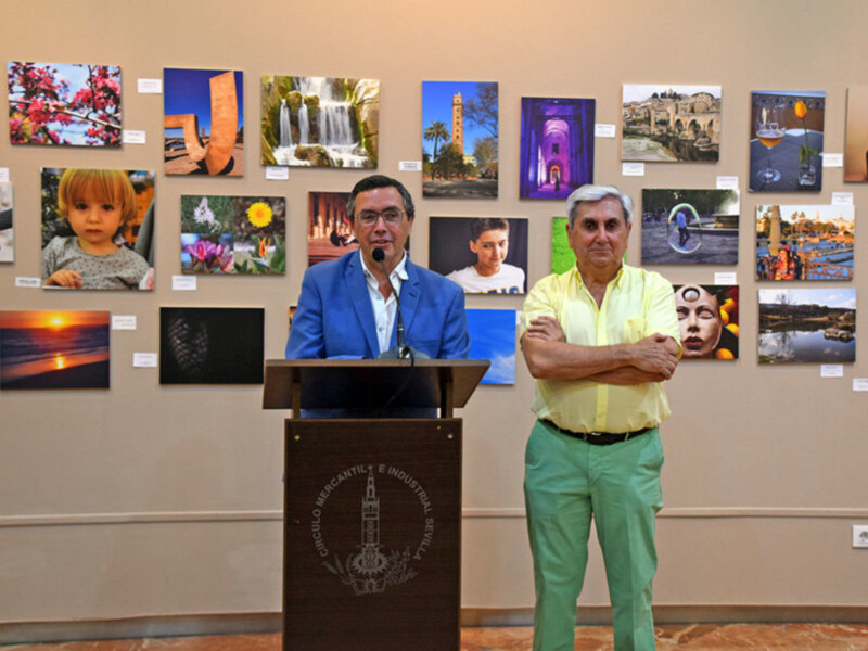 Dos hombres en un evento, uno en un escenario con un micrófono y el otro con las manos cruzadas frente a una pared con varias fotos expuestas.