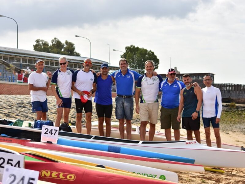 Equipo de remo posando junto a sus botes en la orilla del río.
