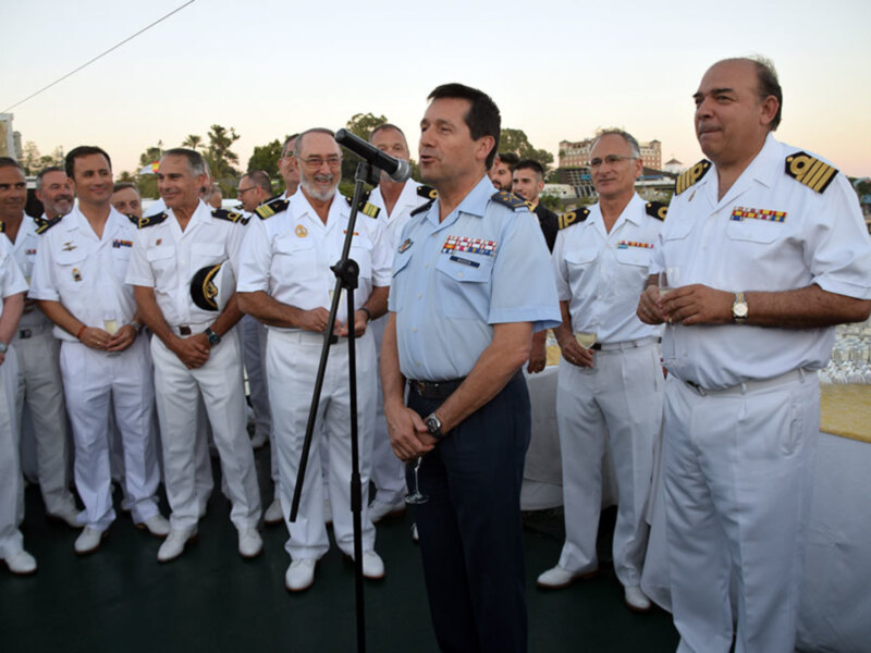 Un oficial de la marina habla en un evento con otros oficiales uniformados en un entorno acuático al atardecer.
