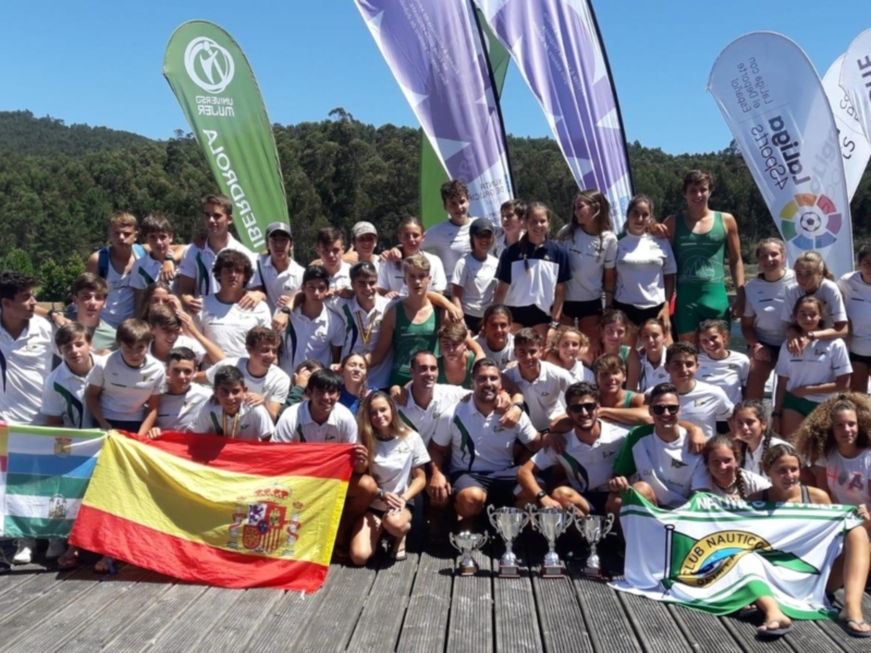 Equipo de remo celebrando con banderas y trofeos en un muelle junto al agua.