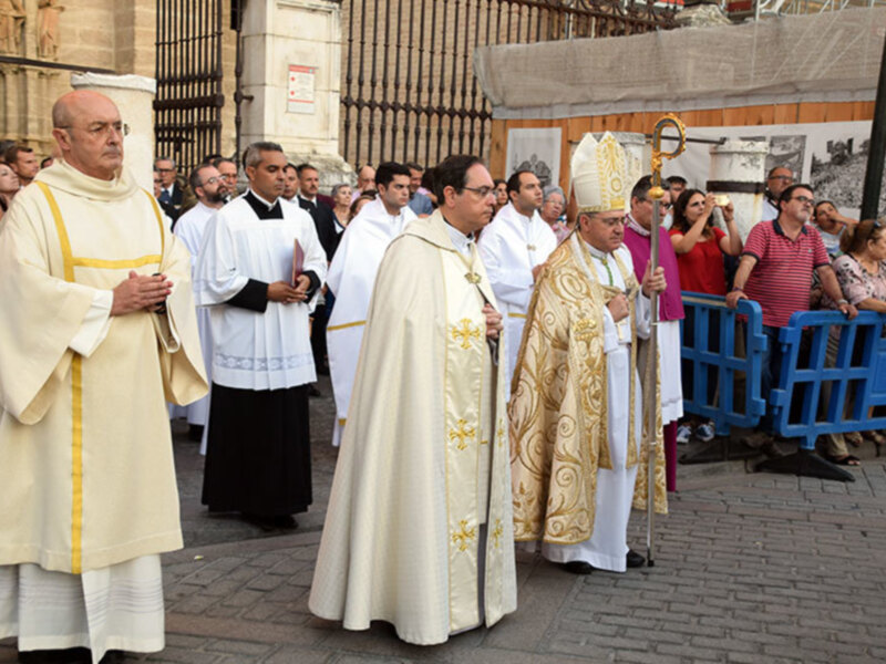 Procesión religiosa con sacerdotes y fieles frente a una catedral.