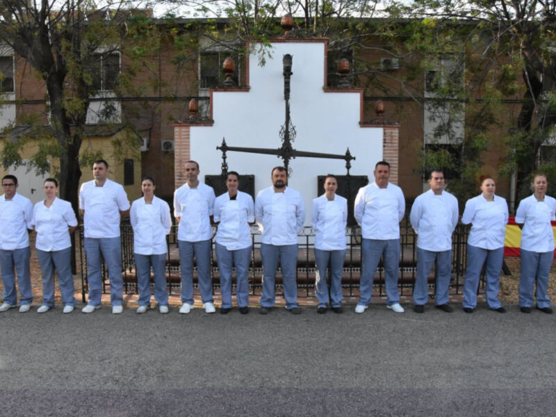 Equipo de cocina en uniforme blanco frente a una fachada de edificio histórico, con una bandera española en el fondo.