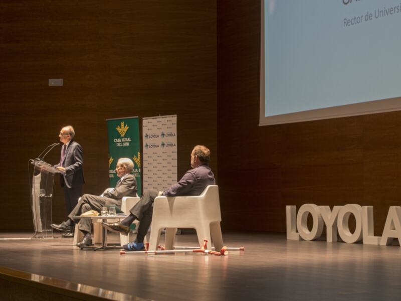 Conferencia en Loyola con el Rector de la Universidad Loyola y otros participantes en un escenario bien iluminado.