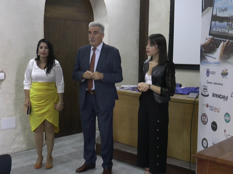 A tres personas en un entorno formal, posiblemente una conferencia o presentación. La persona central es un hombre con traje y corbata, mientras que las dos personas a su izquierda son una mujer en un vestido blanco y falda amarilla, y otra con blusa negra. En el fondo hay un póster informativo sobre eventos culturales y educativos.