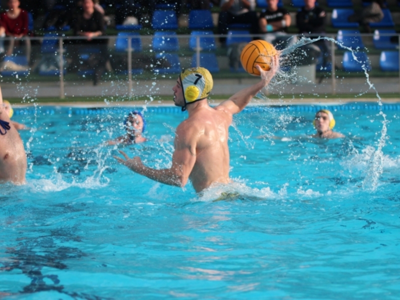 Jugadores de waterpolo en acción durante un partido, con balón en el aire y espectadores en la grada al fondo.