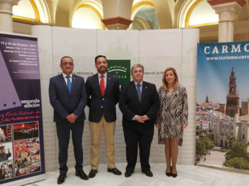 Tres personas posan frente a un panel con imágenes de Carmona y la inscripción 'FECOCAR segunda edición de Carmona'.