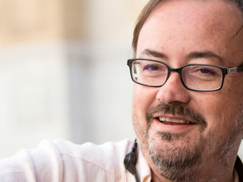 Retrato de hombre con barba y gafas sonriendo, fondo suave y neutro.