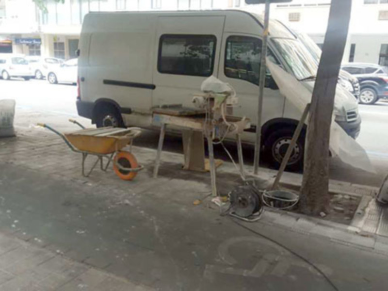 Un carril de trabajo con una carretilla y una mesa de trabajo en la calle. En el fondo, se puede ver un vehículo blanco estacionado en la acera. La escena sugiere que se está trabajando en la calle o realizando mantenimiento en la vía pública.