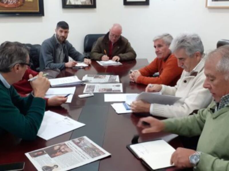 Un grupo de personas se reúne en una sala de conferencias, sentadas alrededor de un escritorio con documentos y portátiles. La imagen muestra una reunión formal, posiblemente de trabajo o negociación.