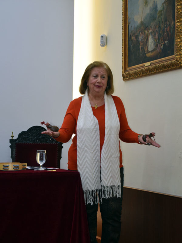 Una mujer con una bufanda blanca y una camisa roja, posando con las manos extendidas en una sala con una mesa roja y un cuadro en el fondo.