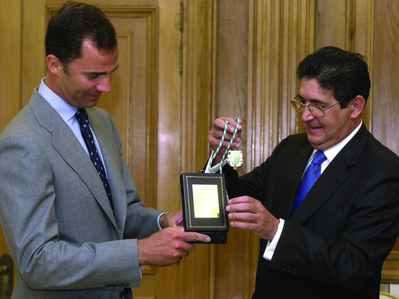 Un hombre en traje gris entrega una medalla a otro hombre en traje negro, ambos sonríen y miran el trofeo.