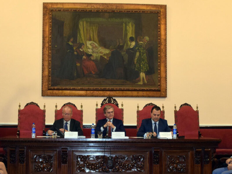 Reunión de autoridades en un salón con pintura histórica en el fondo.