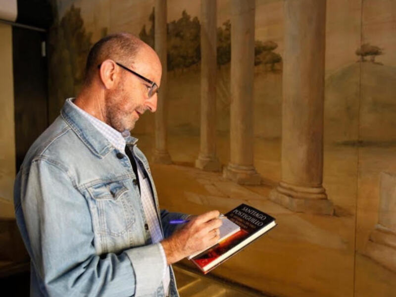 Un hombre examina un libro titulado 'Santiago Portugués' en un entorno arquitectónico clásico.