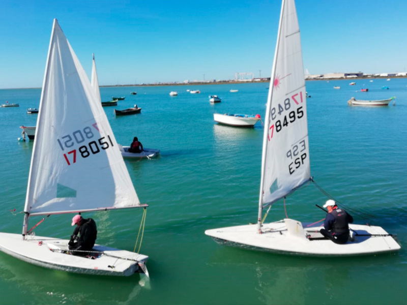 Competición de vela en barcos de vela ligera en el mar, con barcos a la espera en el fondo.