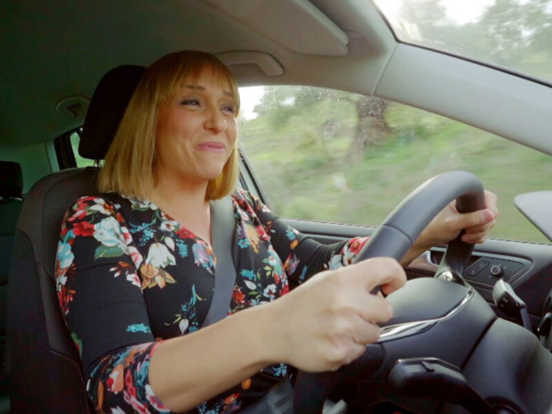 Una mujer con cabello corto y flequillo, vestida con una camisa floral, conduce un automóvil en una carretera con árboles verdes en el fondo.