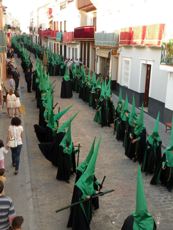 Procession of hooded figures in green cloaks and black robes marching down a narrow street, flanked by onlookers and buildings with balconies.