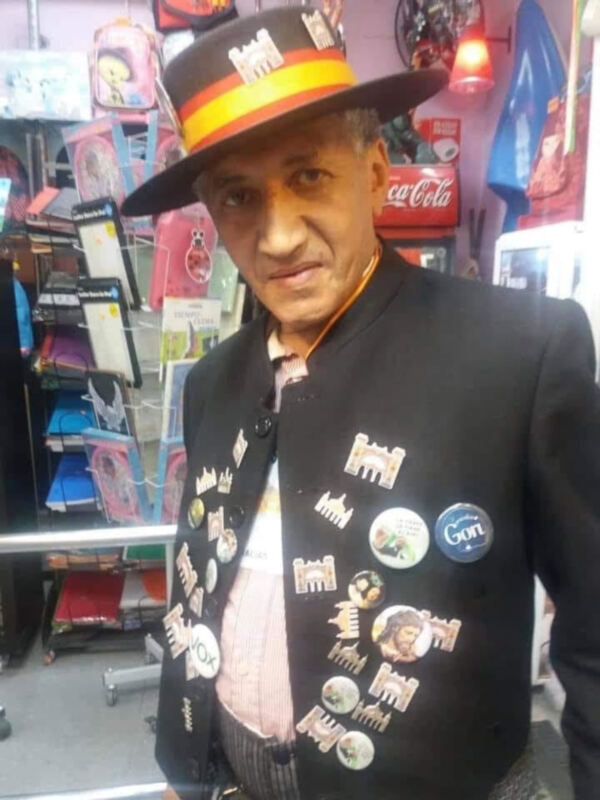 Hombre con sombrero y chaleco lleno de insignias y botones, posando en una tienda de recuerdos.