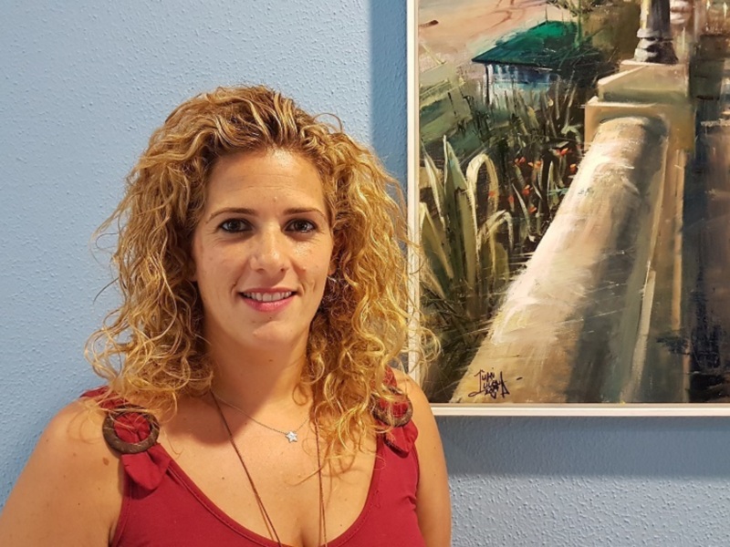 A una mujer con cabello rizado rubio, vestida con un top rojo y una cadena colgante. A su lado derecha hay una pintura que representa un puente con vegetación y edificios en el fondo. La pared de fondo es azul.