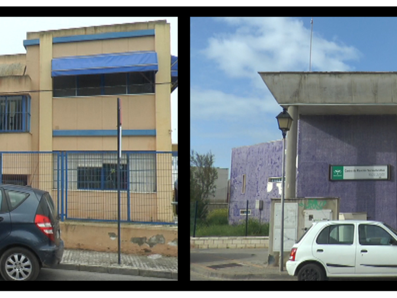 Dos edificios industriales con azulejos y ventanas. En la calle, dos coches estacionados frente a los edificios. El cielo está despejado con algunas nubes.