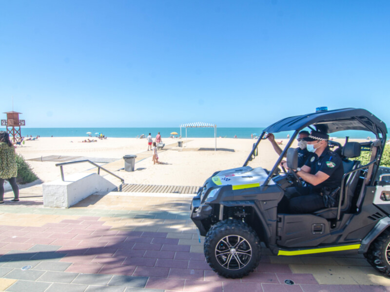 Un policía en un vehículo todoterreno patrulla la playa, con gente disfrutando del sol y el mar en el fondo. La imagen muestra un entorno tranquilo y seguro, con un cielo azul claro y una arena blanca.