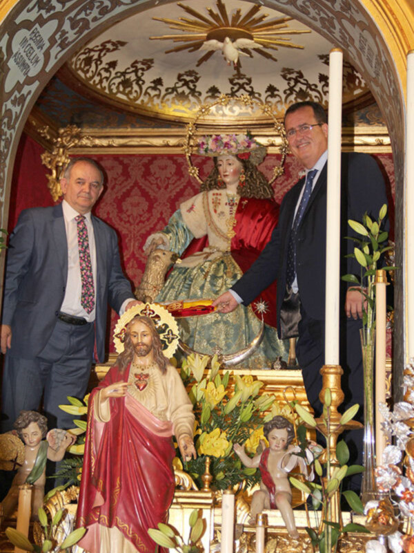 Dos hombres en trajes posan frente a una imagen religiosa en un altar ornamental, rodeado de flores y candelabros.