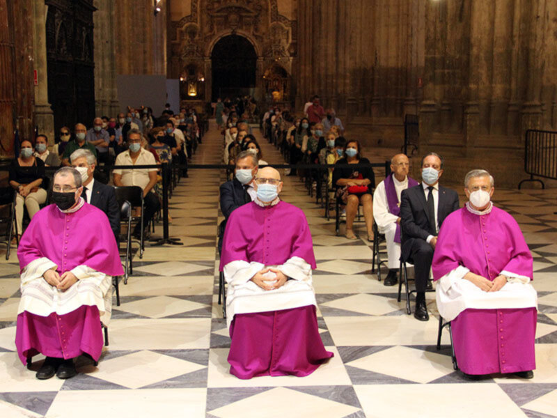 Una ceremonia religiosa en una catedral con varios sacerdotes vestidos de rojo y blanco, sentados en filas. Todos llevan máscaras faciales para prevenir el COVID-19.