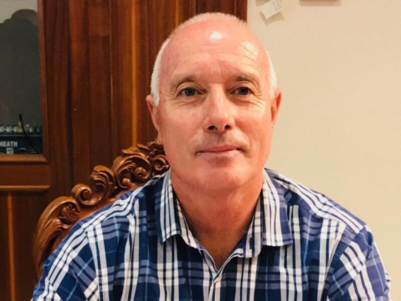 Un hombre con cabello blanco y una camisa a cuadros azul y blanco, sentado en una silla de estilo clásico con detalles dorados, frente a un fondo de madera y una pared blanca.