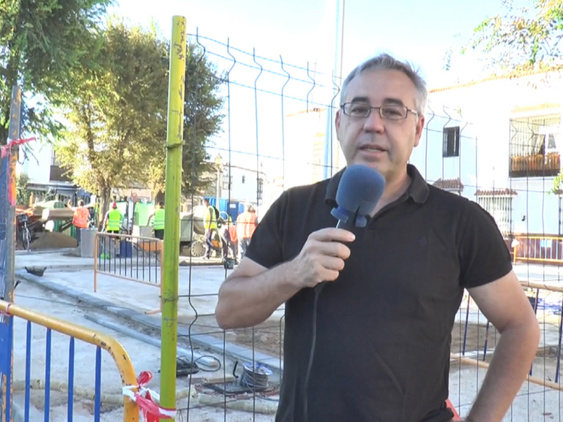 Reportero en una zona de obras, sosteniendo un micrófono. Fondo con trabajadores y estructuras de construcción.