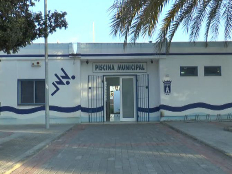 Piscina Municipal con su nombre en letras grandes y un escudo en la puerta principal, rodeada de árboles y palmeras.