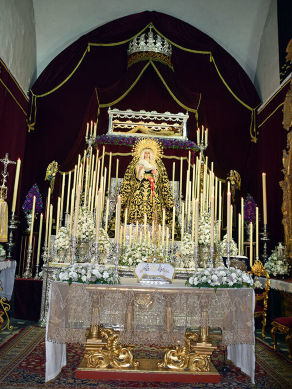 Altar con una imagen religiosa en el centro, rodeada de flores y velas, con una velada y un manto dorado en el fondo.