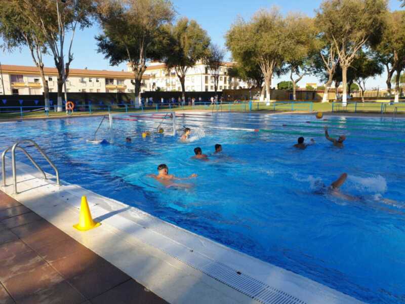 Jóvenes jugando waterpolo en una piscina al aire libre con árboles y edificios al fondo.