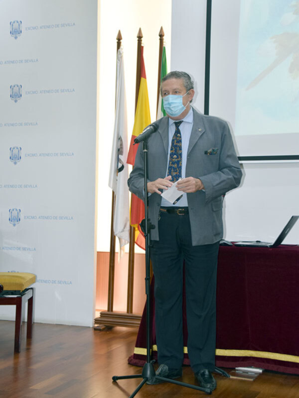 Persona hablando en un evento del Ateneo de Sevilla, con micrófono y banderas en el fondo.