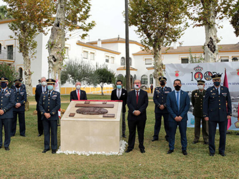 Celebración de un evento militar en un parque, con oficiales y civiles en uniforme, frente a un monumento con placas de madera. El evento parece ser un homenaje o conmemoración, con la presencia de personalidades importantes y un ambiente formal.