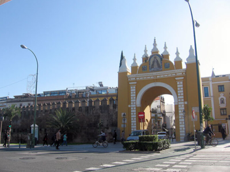 Puerta monumental amarilla con detalles blancos, frente a un edificio histórico. Personas caminan por la calle y ciclistas pasan cerca de la entrada.