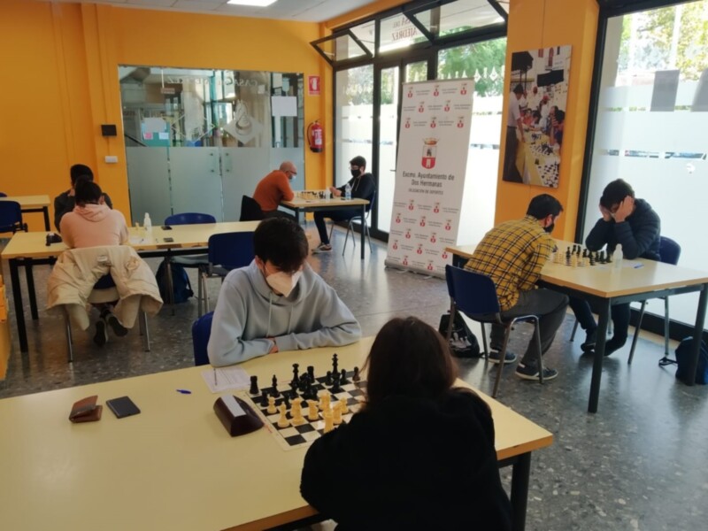 Jóvenes jugando al ajedrez en un salón escolar con mesas y tableros de ajedrez en el centro.