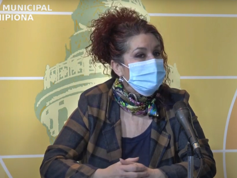 Una mujer habla en una conferencia de prensa, con una máscara médica y un fondo con un mapa de la ciudad.