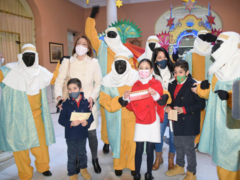 Una familia feliz con niños y adultos vestidos de personajes navideños, posando frente a un arco decorado con estrellas y una figura de Navidad. El ambiente es festivo, con luces navideñas en el fondo y una decoración navideña en la pared.