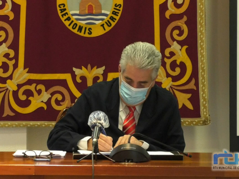 Un hombre se dirige a un público mediante un micrófono, con una bandera de fondo que contiene el texto 'CAEPIONISIS TURRIS'.