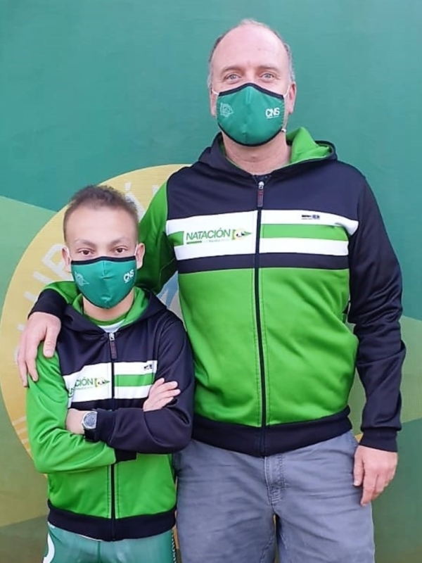 Un padre y su hijo, ambos con chaquetas verdes y máscaras de natación, posan frente a un fondo verde con un logo de natación.