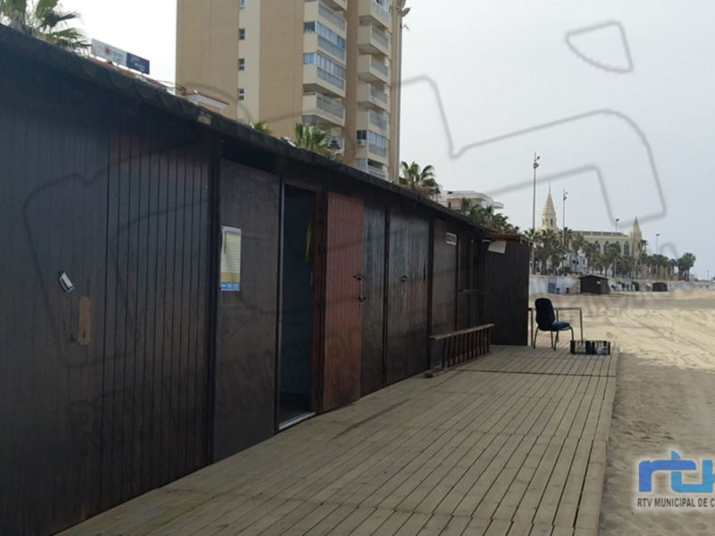 Un pequeño toldo de madera en la playa, con una silla y una mesa a su lado, frente a un edificio de varios pisos y la torre de la iglesia de la ciudad en el fondo.